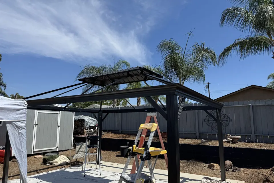 Hardtop gazebo frame mid-build with a ladder in a backyard